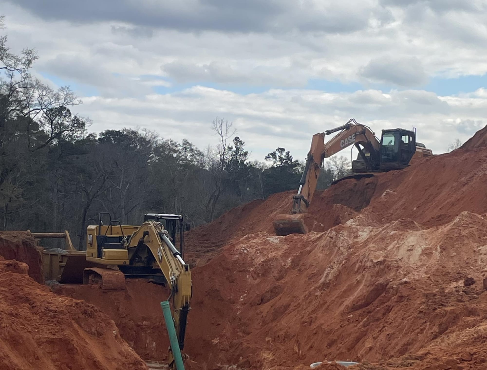 Two excavators working a deep red clay cut with a pipe riser in the trench